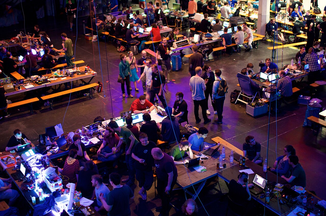 Blick in das Hackcenter der Gulaschprogrammiernacht in Karlsruhe: lange Tischreihen mit Notebooks, Kabeln und Bildschirmen.